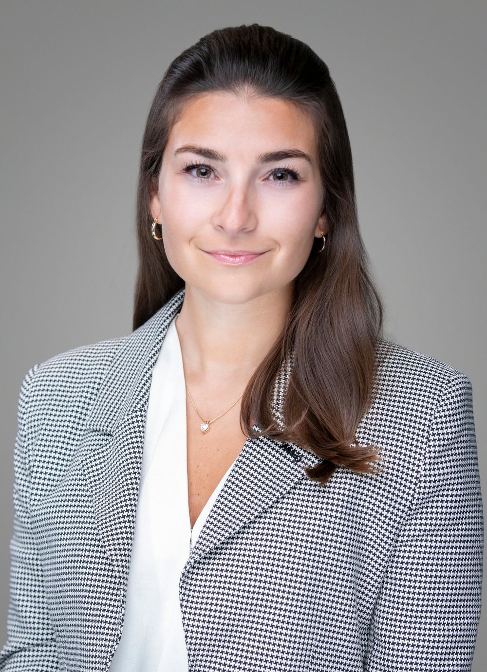 Professional headshot of a young woman in a houndstooth blazer with long brown hair, smiling confidently.