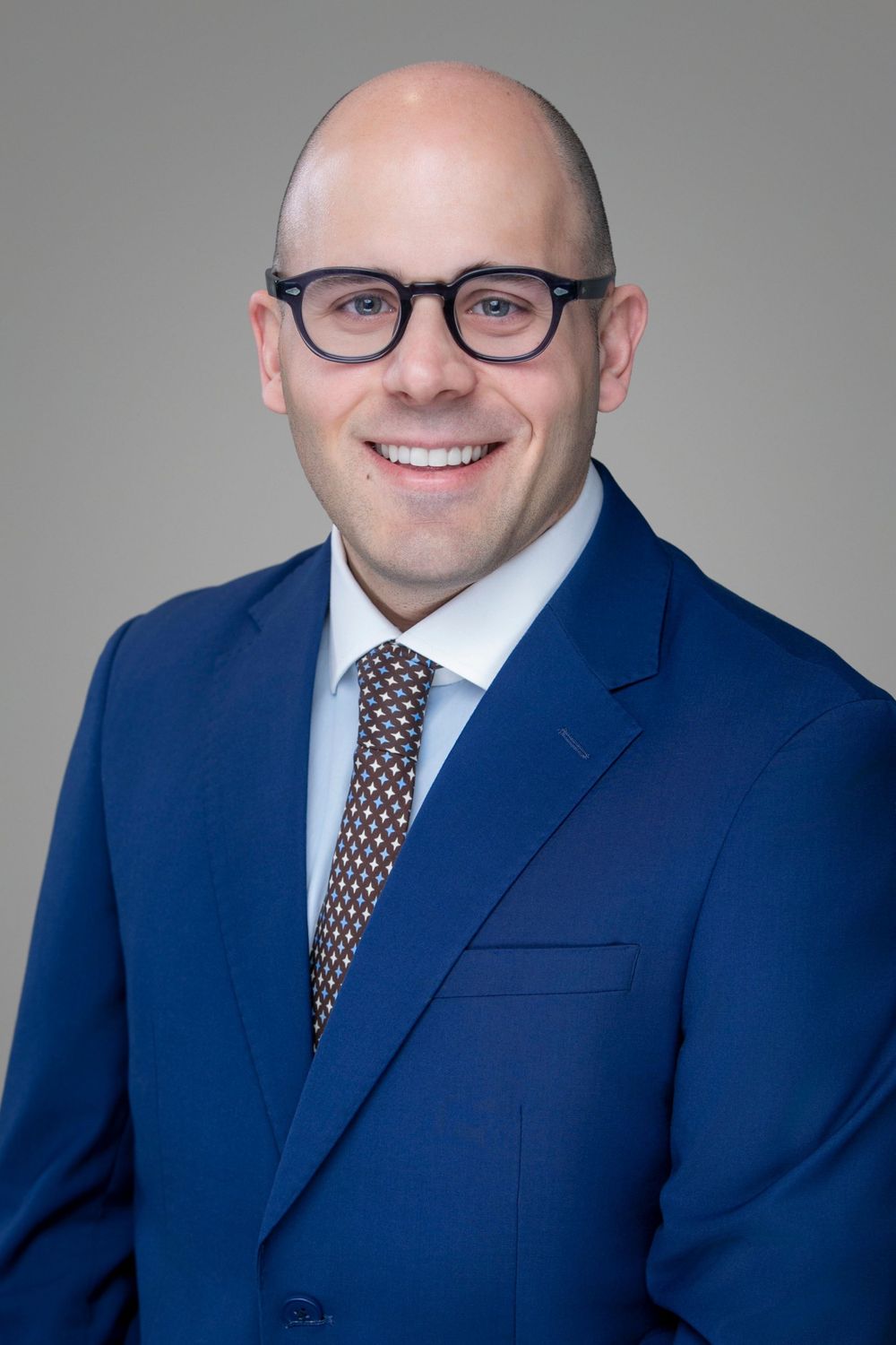 Professional headshot of a smiling bald man wearing glasses, blue suit, and patterned tie against gray background.