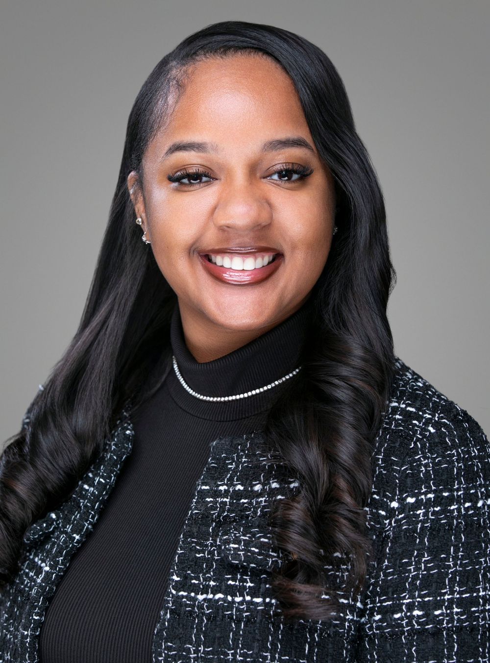 Professional headshot of a smiling young woman in a plaid blazer and black turtleneck with a pearl necklace.
