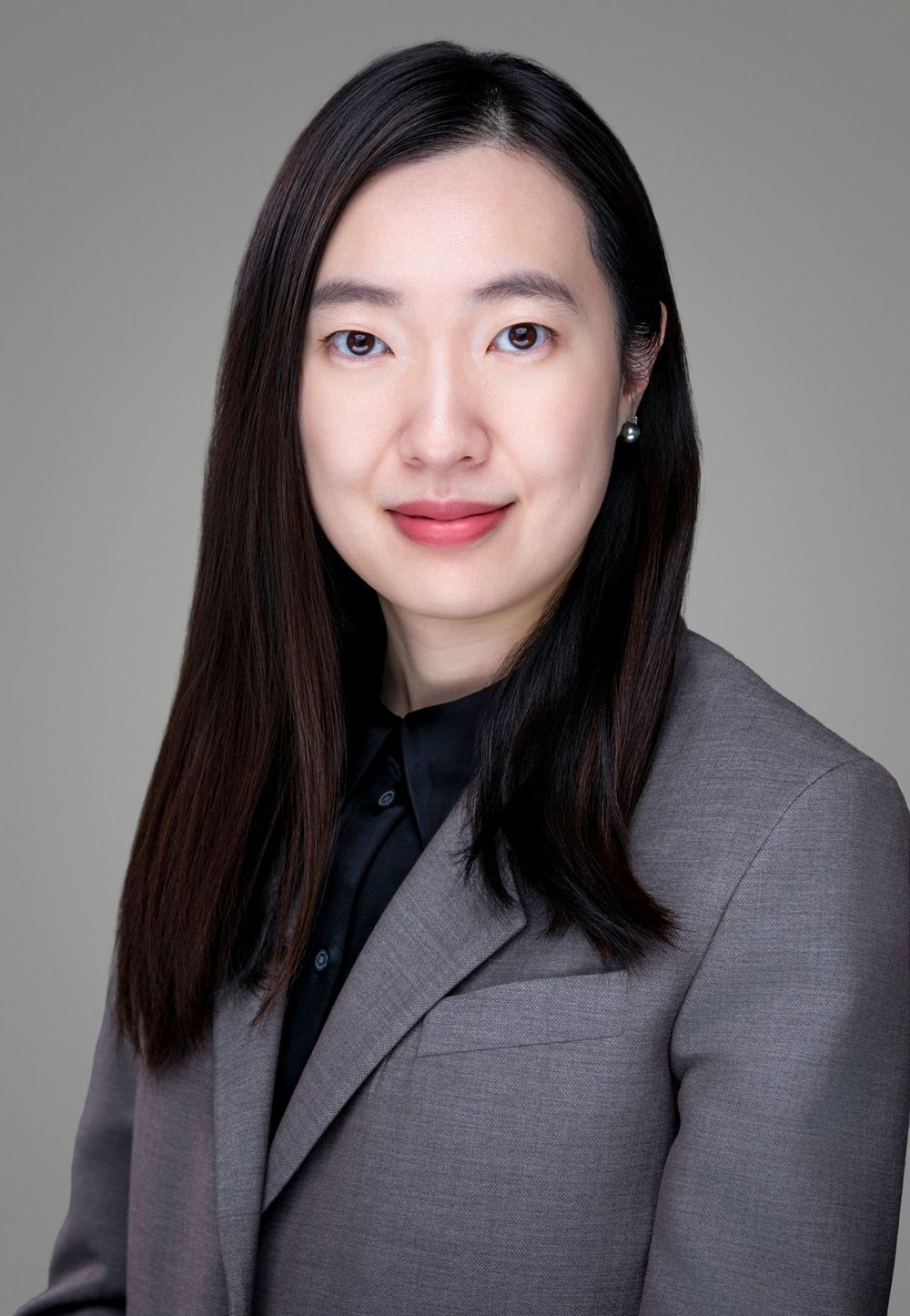 Professional headshot of an Asian woman in a gray blazer with long dark hair, wearing a small earring.