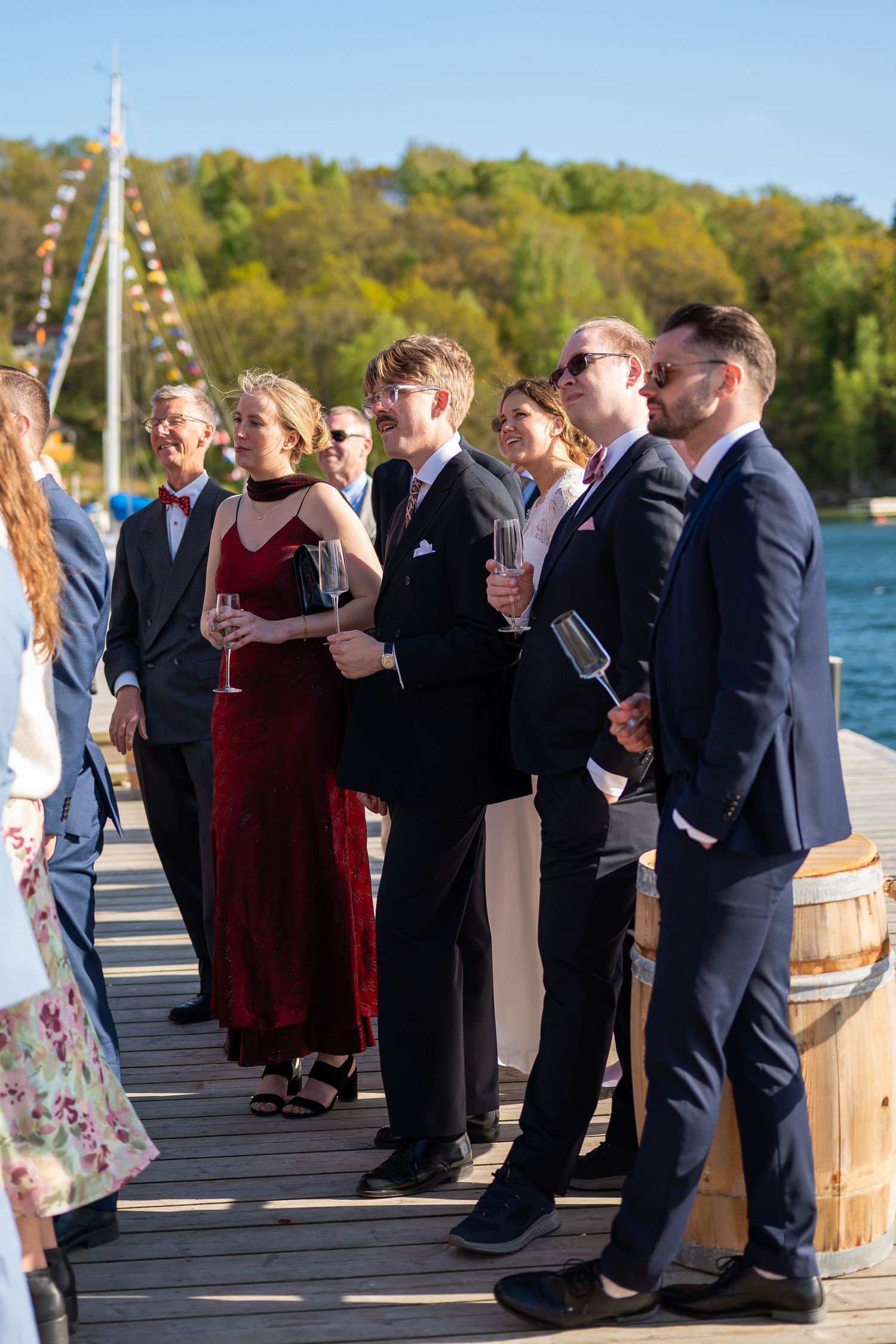 Elegantly dressed wedding guests holding champagne glasses on a wooden dock by a lake on a sunny day.