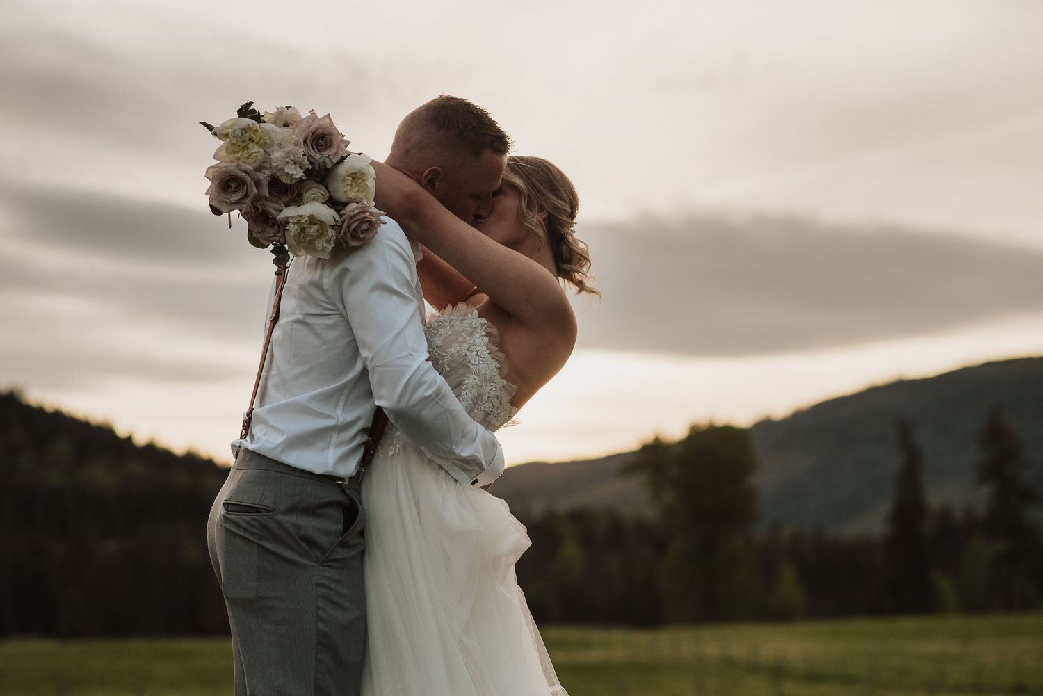 Bride and groom embrace at sunset in a mountain meadow, bride holding white floral bouquet in romantic wedding photo.