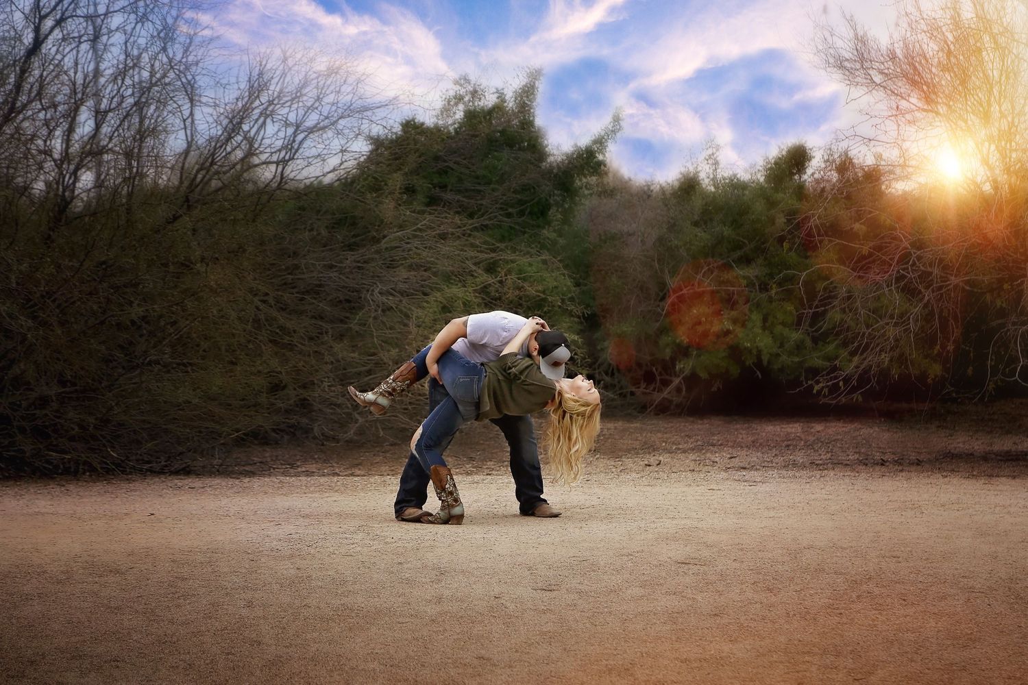 Couple in a romantic dip embrace on a dirt path at sunset, surrounded by autumn trees and a glowing sun flare in the sky.