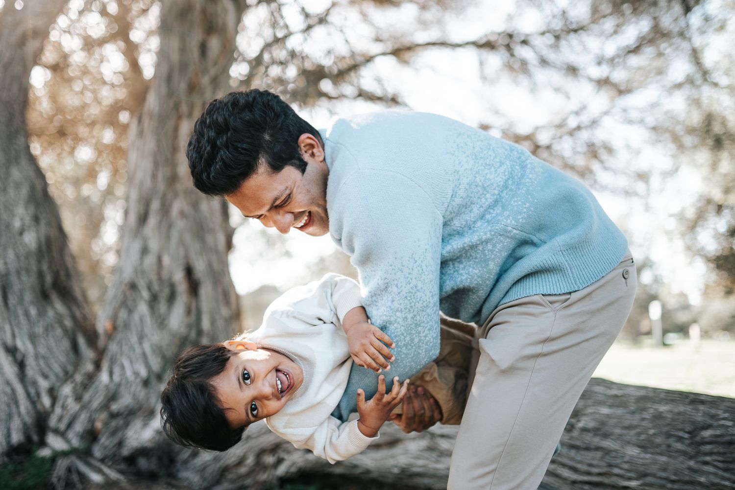 A father playfully lifts his laughing child outdoors near a large tree, sharing a joyful bonding moment during a Melbourne family photoshoot