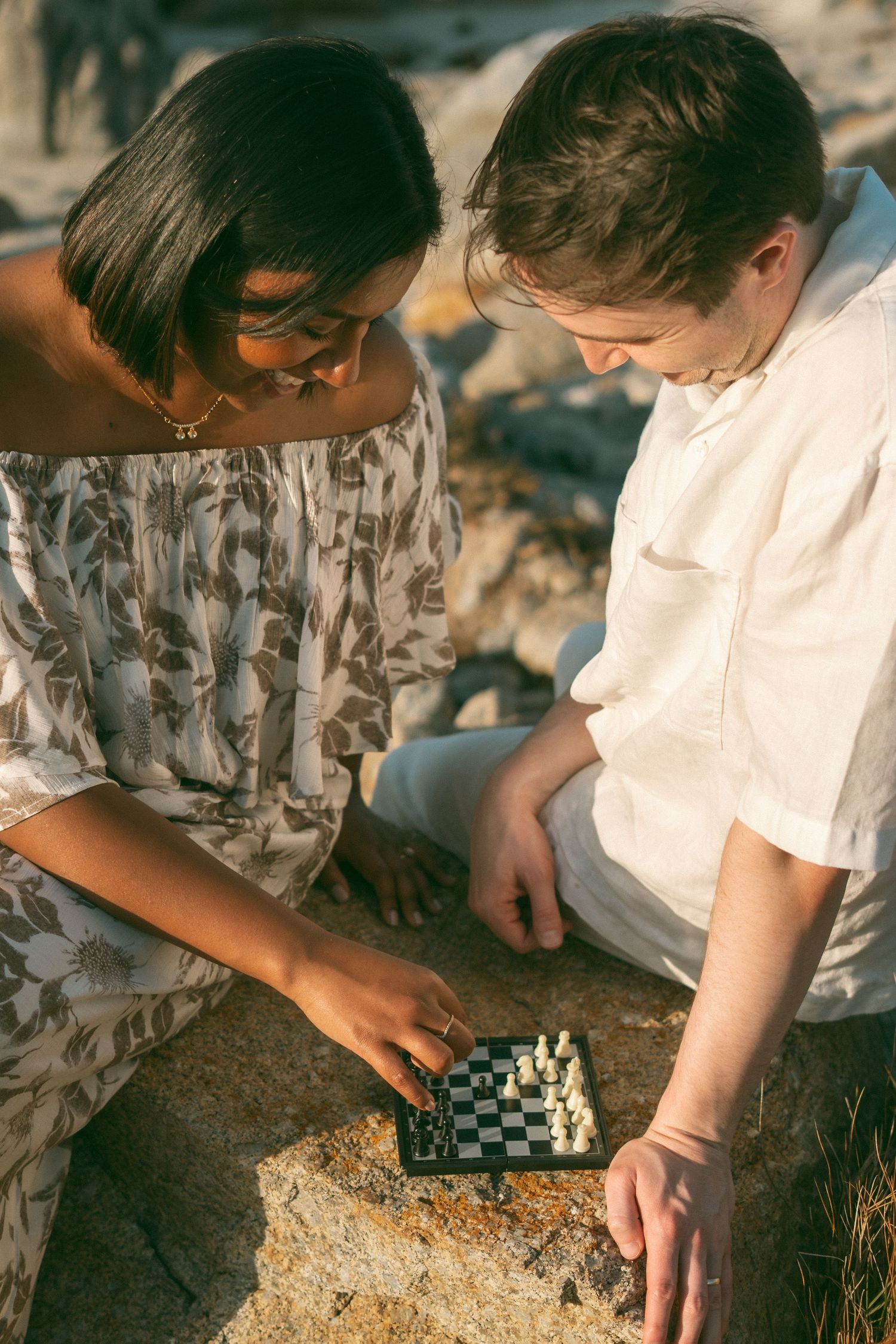 A man and woman sit on rocks outdoors playing a small chess game together in warm golden sunlight.