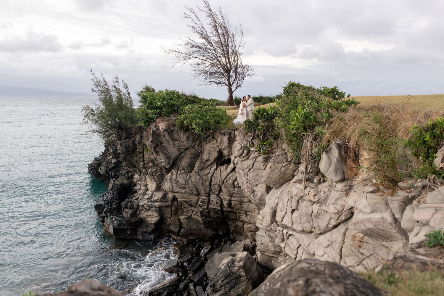Couple stands on dramatic rocky coastal cliffs with lush greenery and ocean views under cloudy skies.
