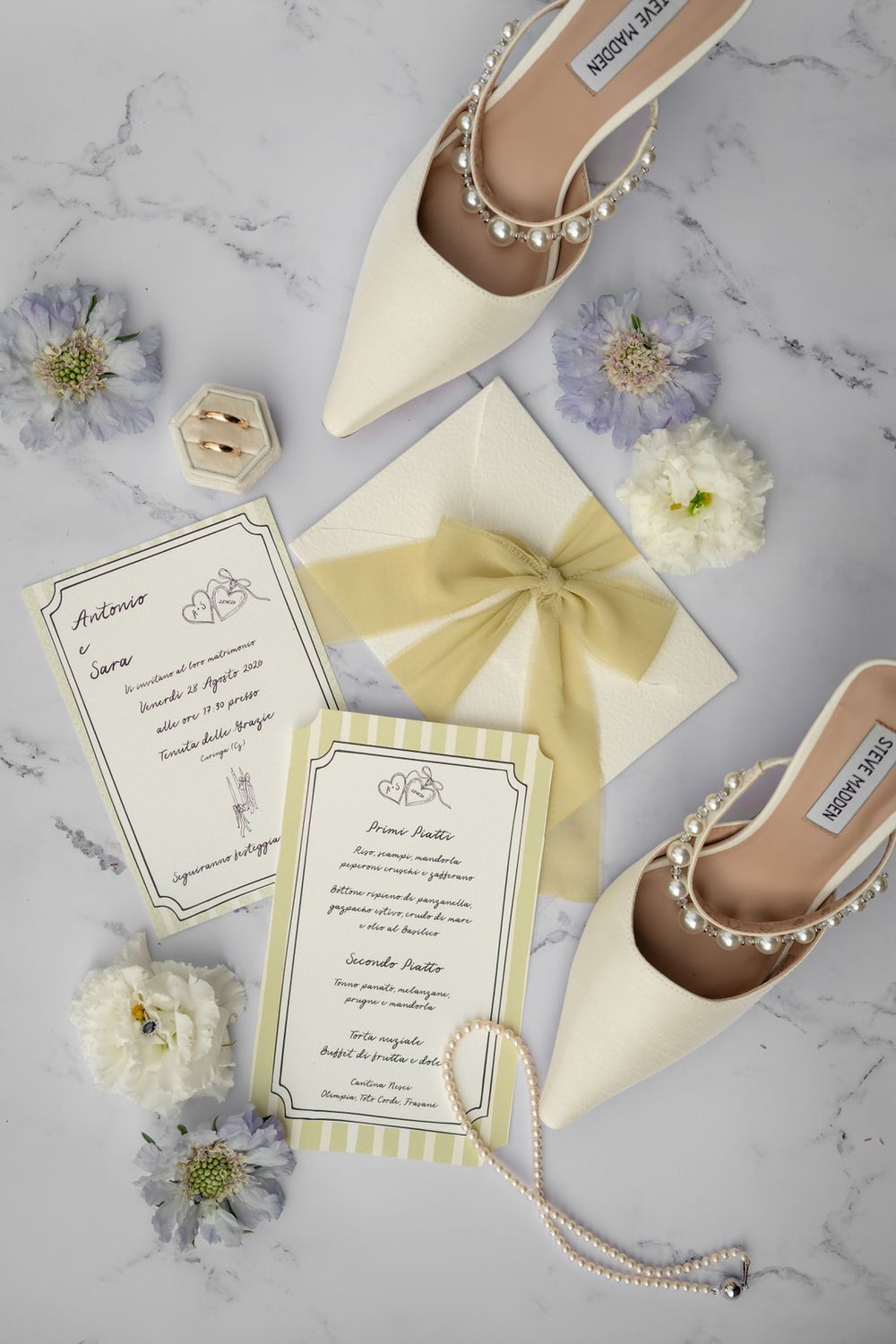 Elegant wedding flat lay with ivory heels, pearl necklace, yellow bow envelope, invitations, and floral accents on marble.