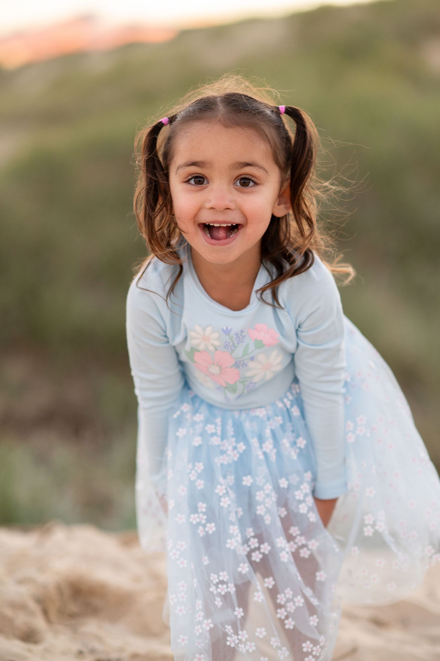 A joyful toddler girl in a light blue floral dress with pigtails smiles outdoors on sandy dunes.