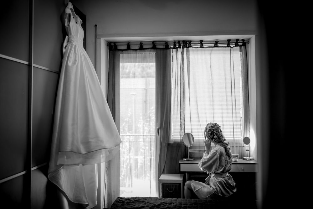 Black and white photo of a bride sitting at a vanity with her wedding dress hanging nearby.