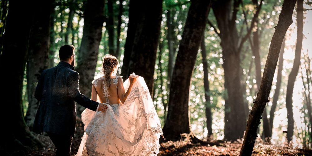 Bride and groom walking hand in hand through a sunlit forest in wedding attire.