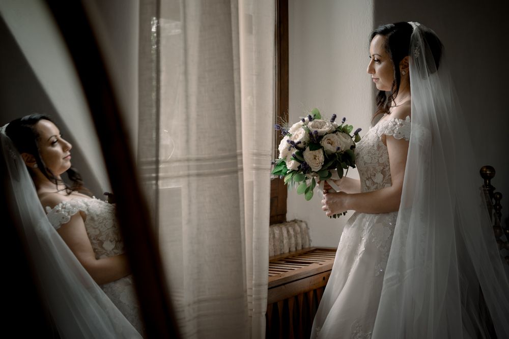 Elegant bride in lace wedding gown holding bouquet reflected in vintage mirror by window.