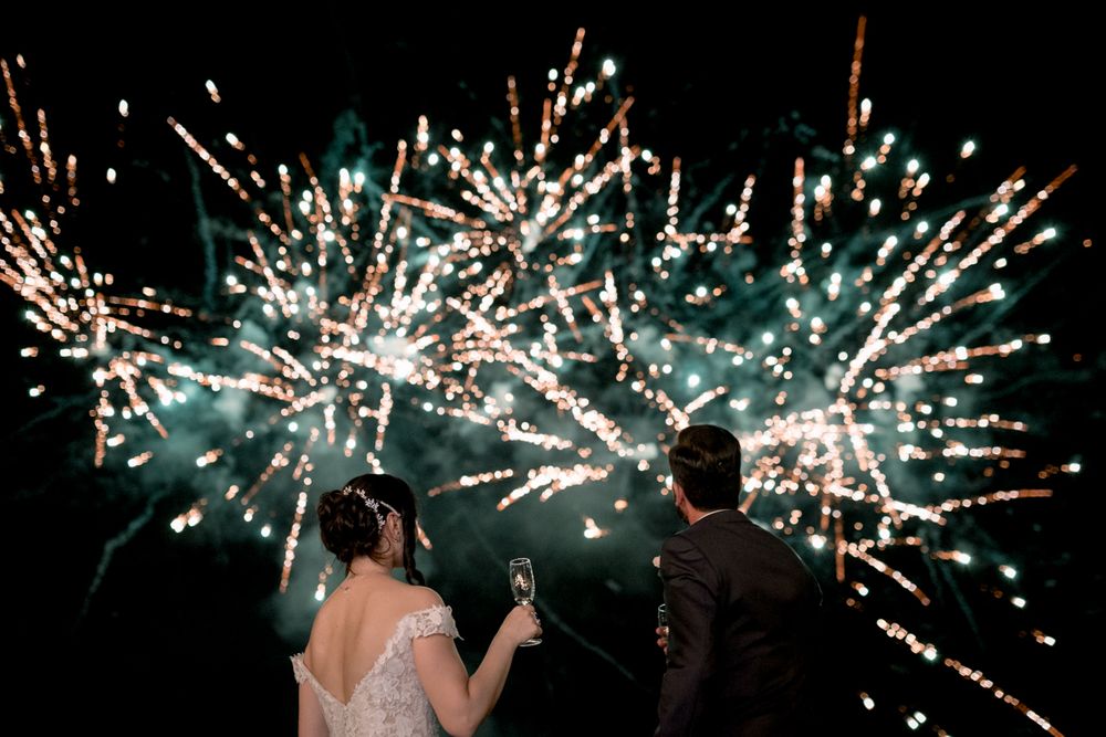 Newlywed couple watching colorful fireworks display at night holding champagne glasses together.