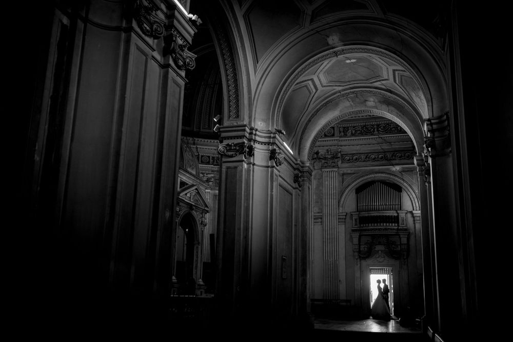 Black and white photo of wedding couple silhouetted in grand cathedral archway with ornate ceiling.