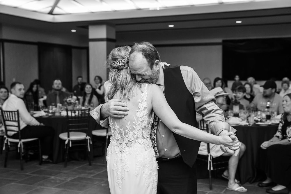 Black and white photo of bride and groom sharing their first dance at wedding reception with guests seated at tables.
