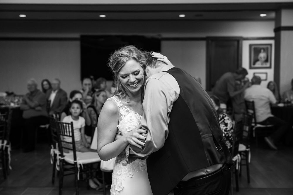 Black and white photo of a bride smiling while sharing a warm hug during a wedding reception.