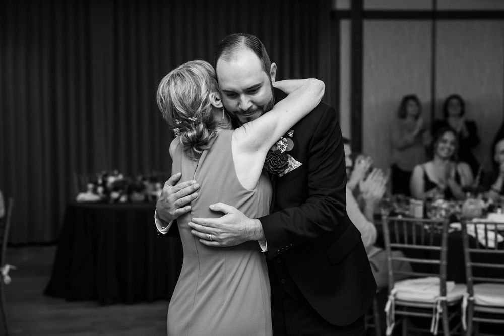 Black and white photo of groom and mother sharing emotional first dance hug at elegant wedding reception.