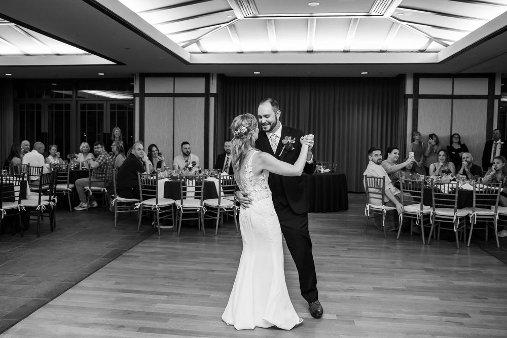 Black and white photo of bride and groom sharing first dance at wedding reception with guests seated at tables.