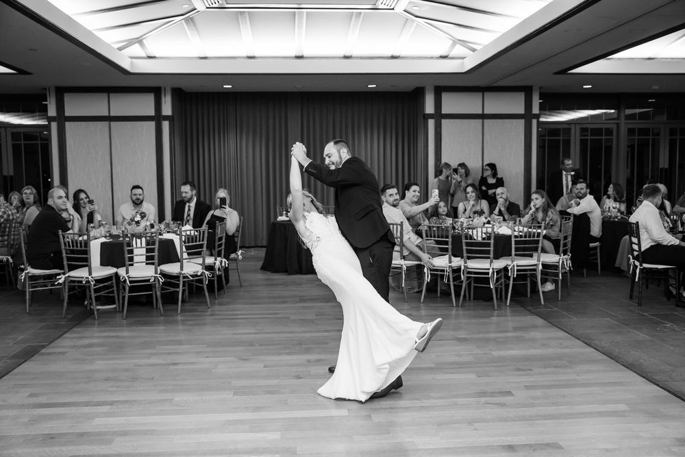 Black and white photo of bride and groom sharing first dance at wedding reception with guests seated at tables.
