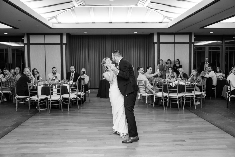 Black and white photo of a couple sharing their first dance at a wedding reception with guests seated at tables.