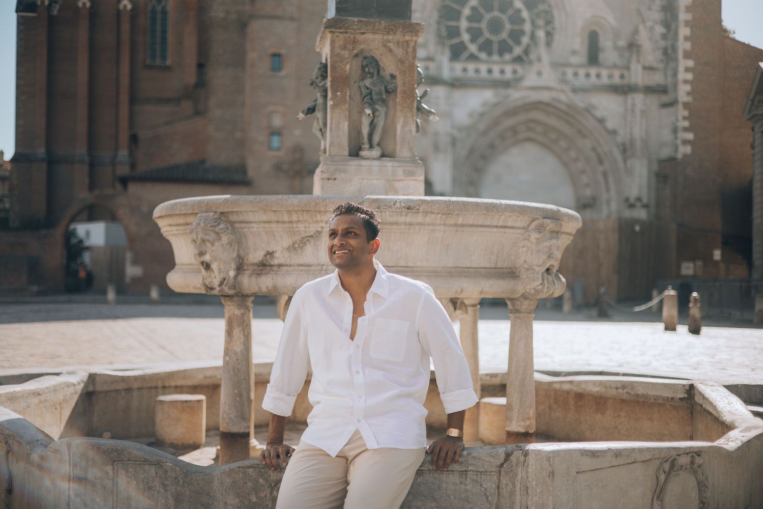 Man in white shirt sitting on ornate stone fountain in sunny European plaza with historic cathedral backdrop.
