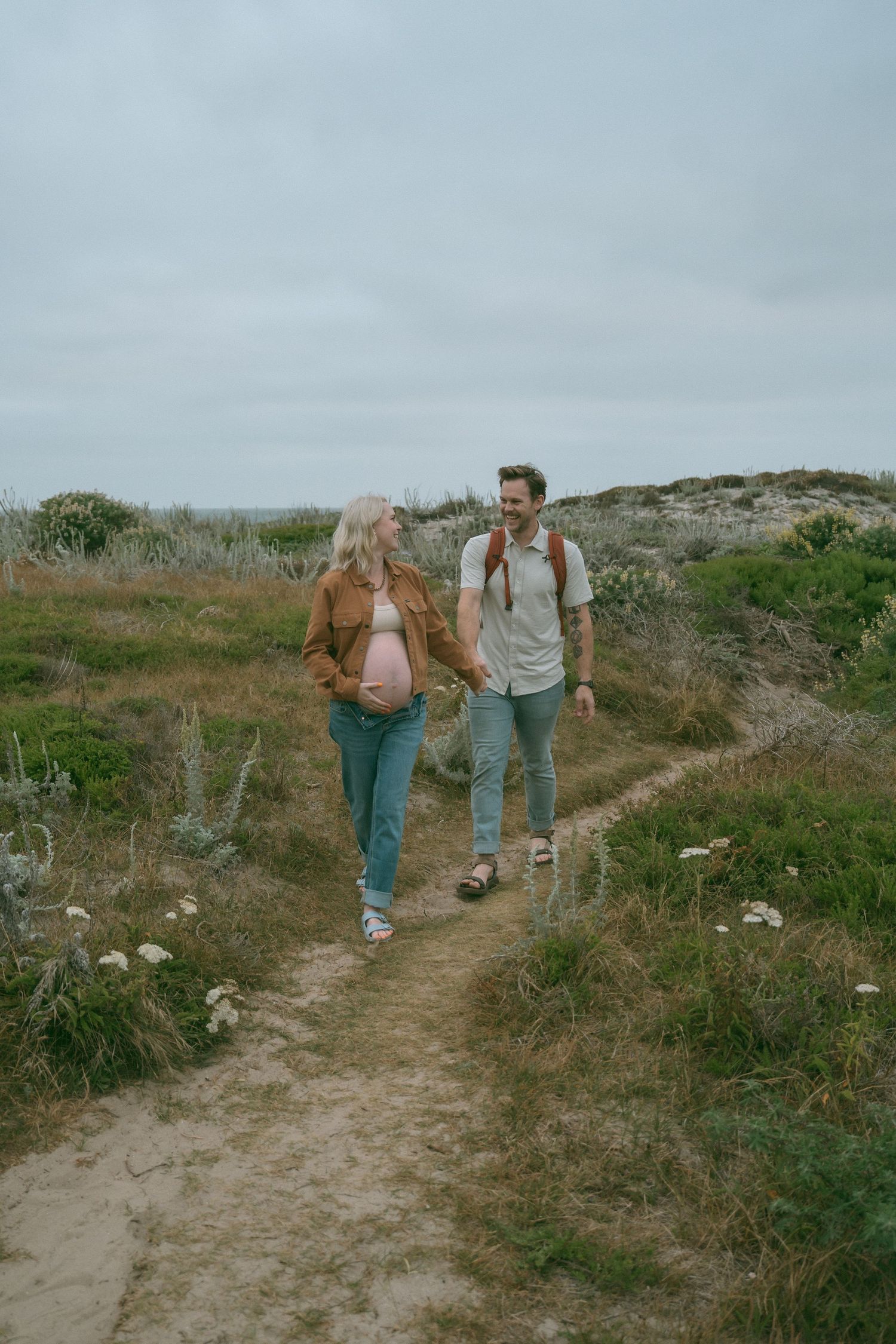 Pregnant couple holding hands while hiking on a scenic coastal trail surrounded by green shrubs under overcast skies.