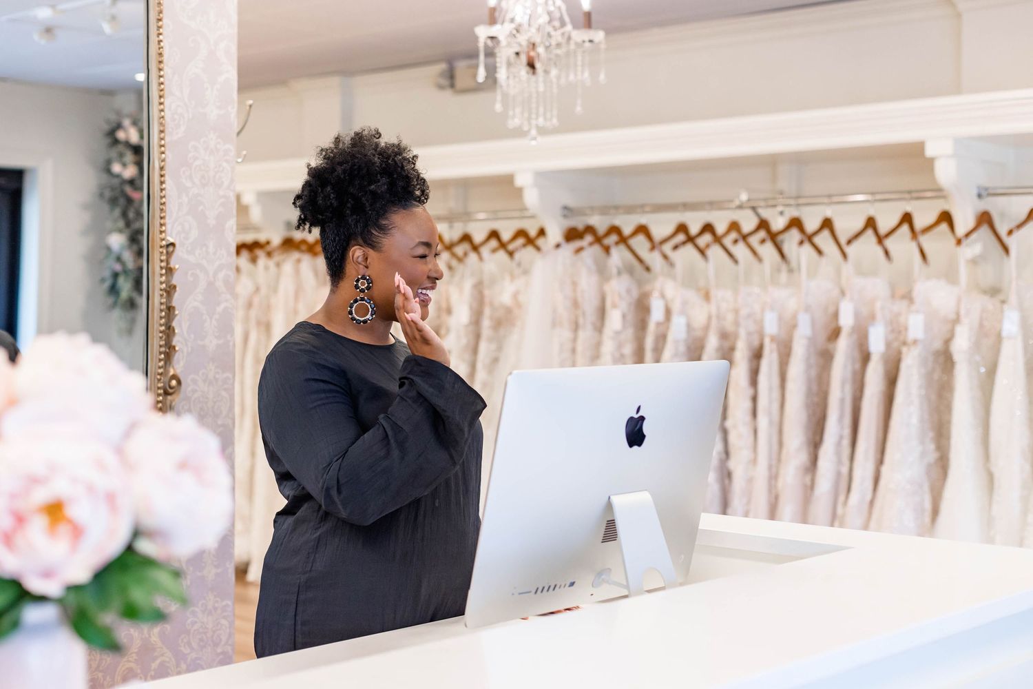 A woman in a black dress works at an Apple iMac computer at a bridal boutique reception desk surrounded by wedding gowns.