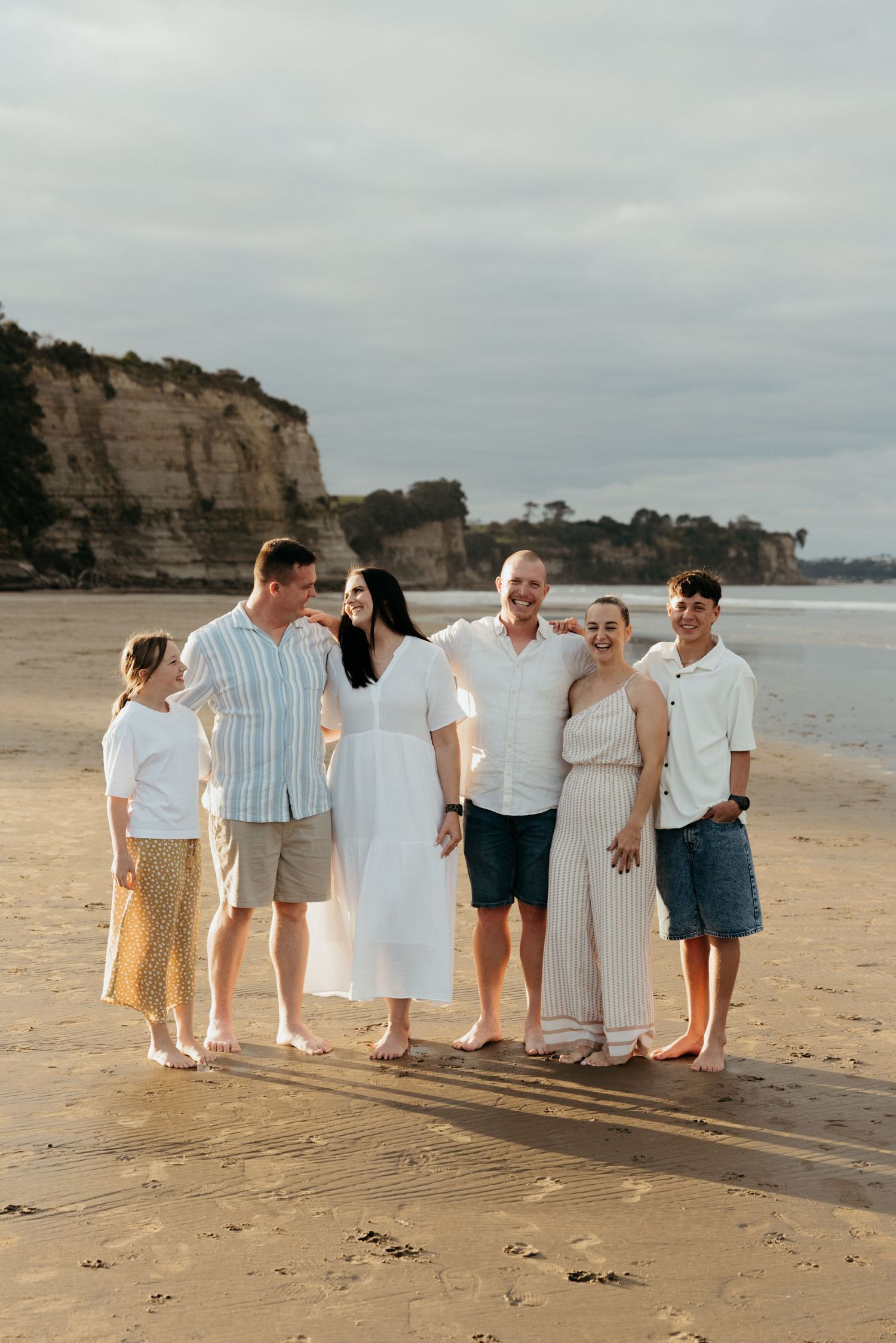 Family of six smiling together on a sandy beach with rocky cliffs in the background at golden hour.