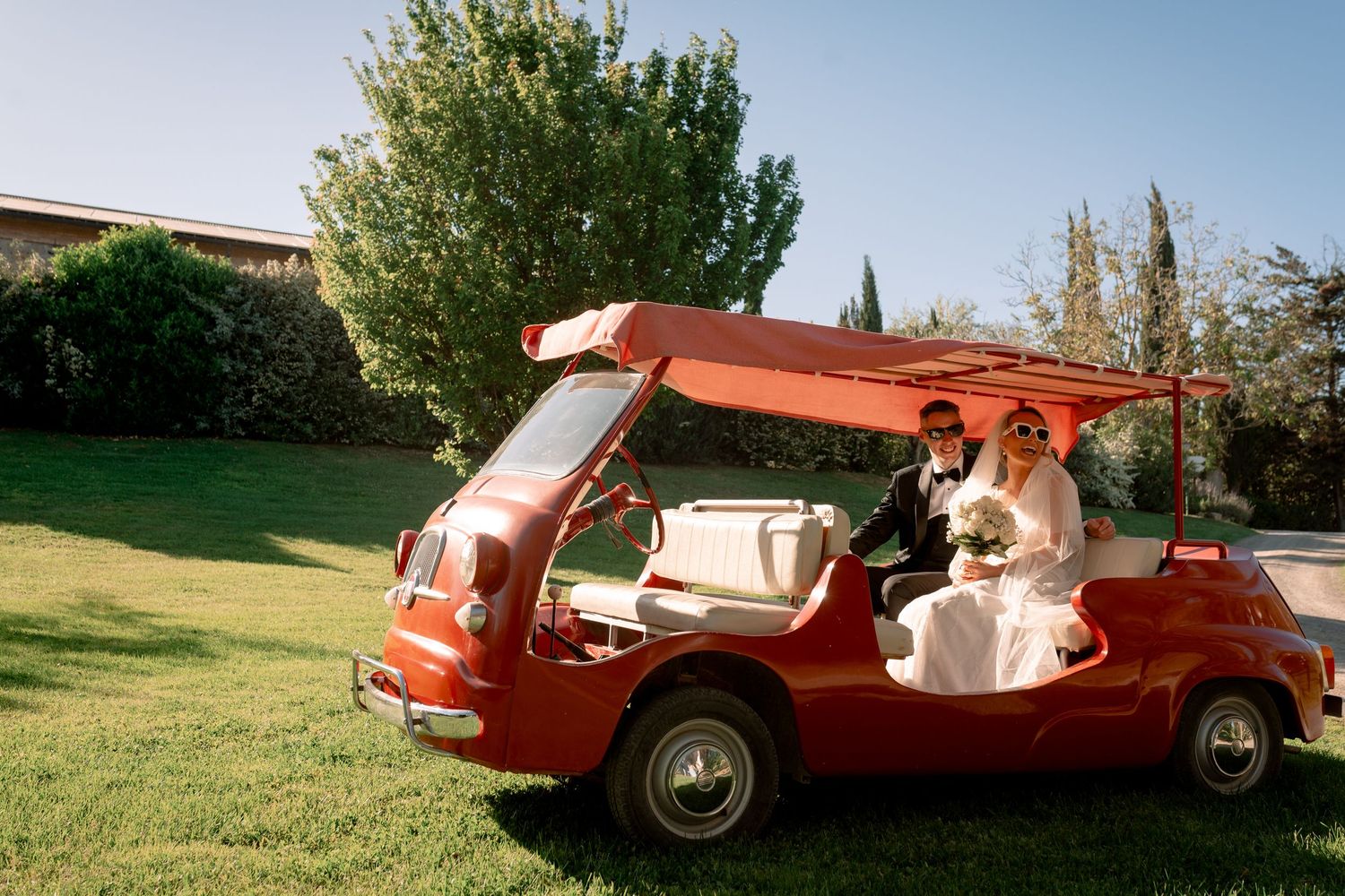 Bride and groom riding in a vintage red open-top car on green lawn at outdoor wedding venue.