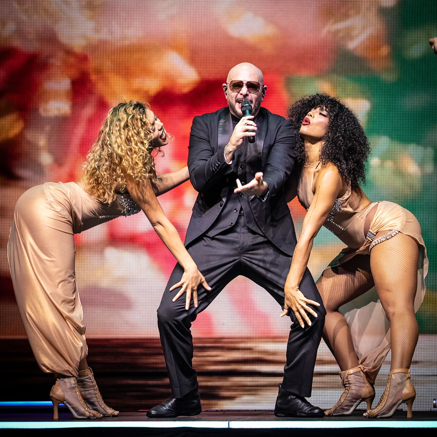 Male performer in black suit sings on stage flanked by two female dancers in nude outfits under colorful lights.