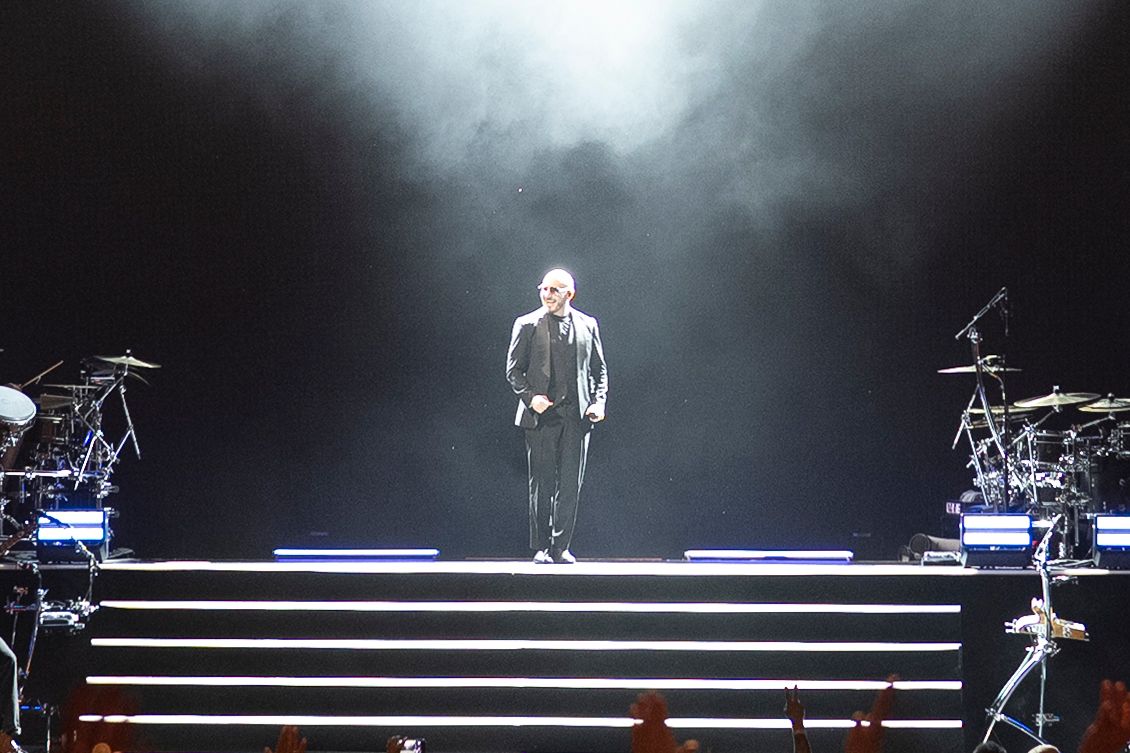 A lone performer stands on a dramatic lit staircase stage with drum kits on either side amid smoky atmosphere.