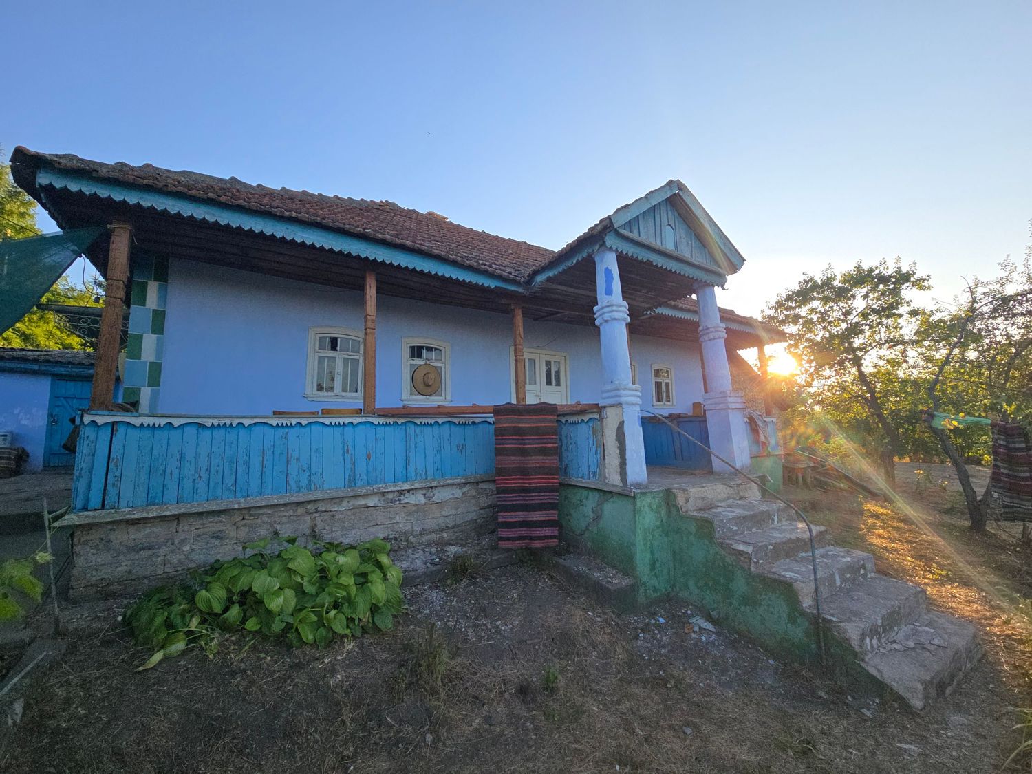 Blue rural house with white columns and porch at sunset, surrounded by trees and garden in a countryside setting.