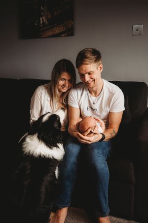A couple sits on a dark couch with their Border Collie dog and small baby.
