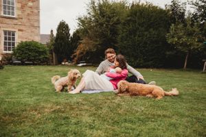 Couple relaxes on grass with two goldendoodle dogs beside a brick building.