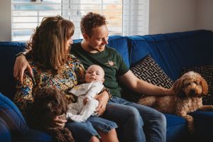 Family cuddles with baby and dogs on blue couch in sunlit living room.