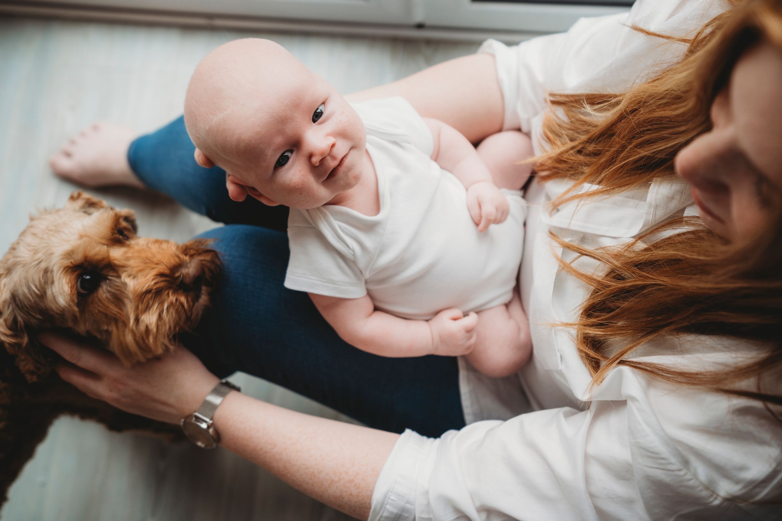 Happy baby in white onesie lying on floor with family and pet dog.