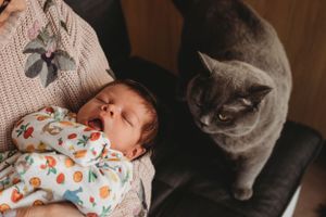 A grey cat sits attentively next to a sleeping baby in a floral onesie.