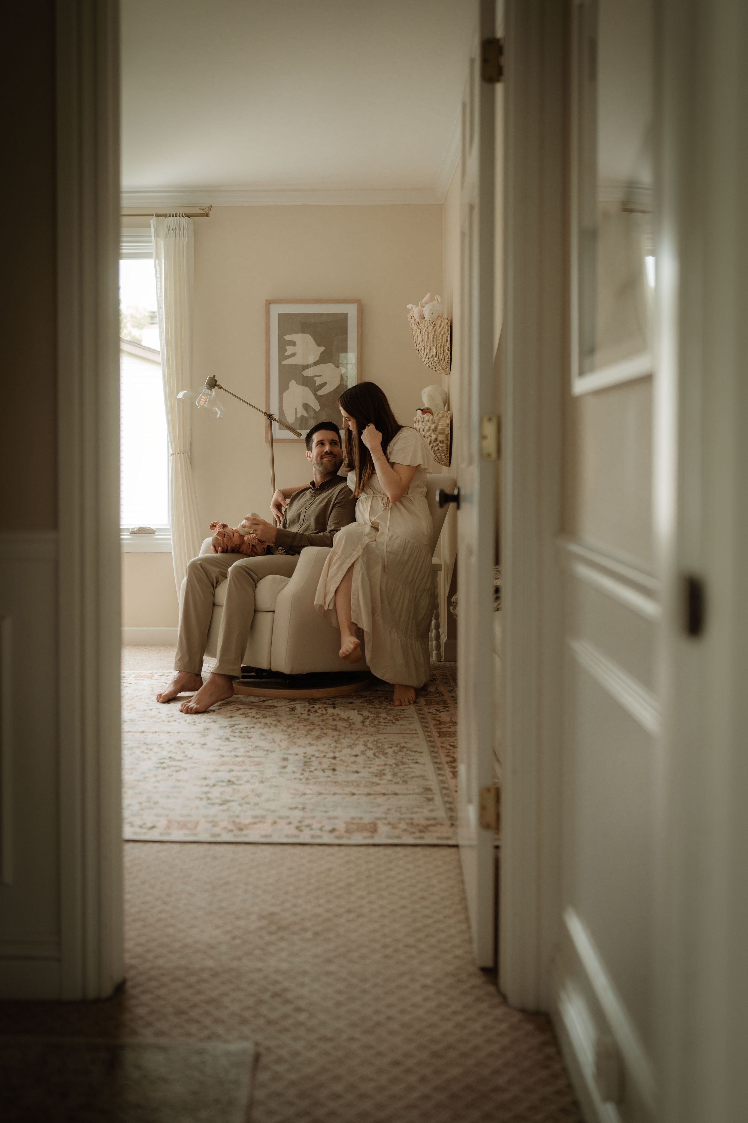 Couple relaxing together in a softly lit living room, seen through a doorway, with a cozy armchair, vintage rug and neutral decor.
