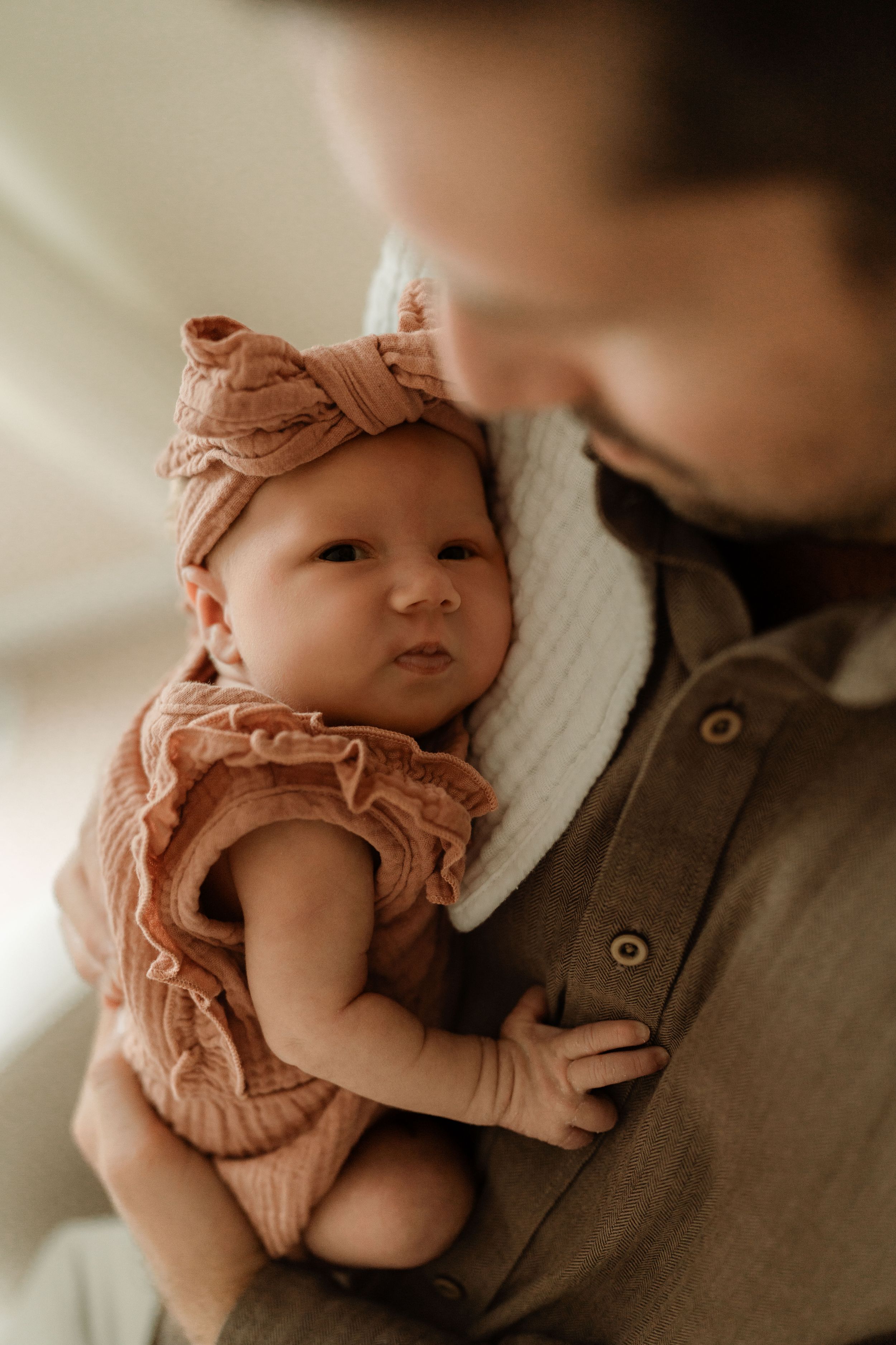 Newborn baby in a dusty rose ruffled romper and knotted headband rests against a man in a button-up shirt with a white muslin burp cloth.