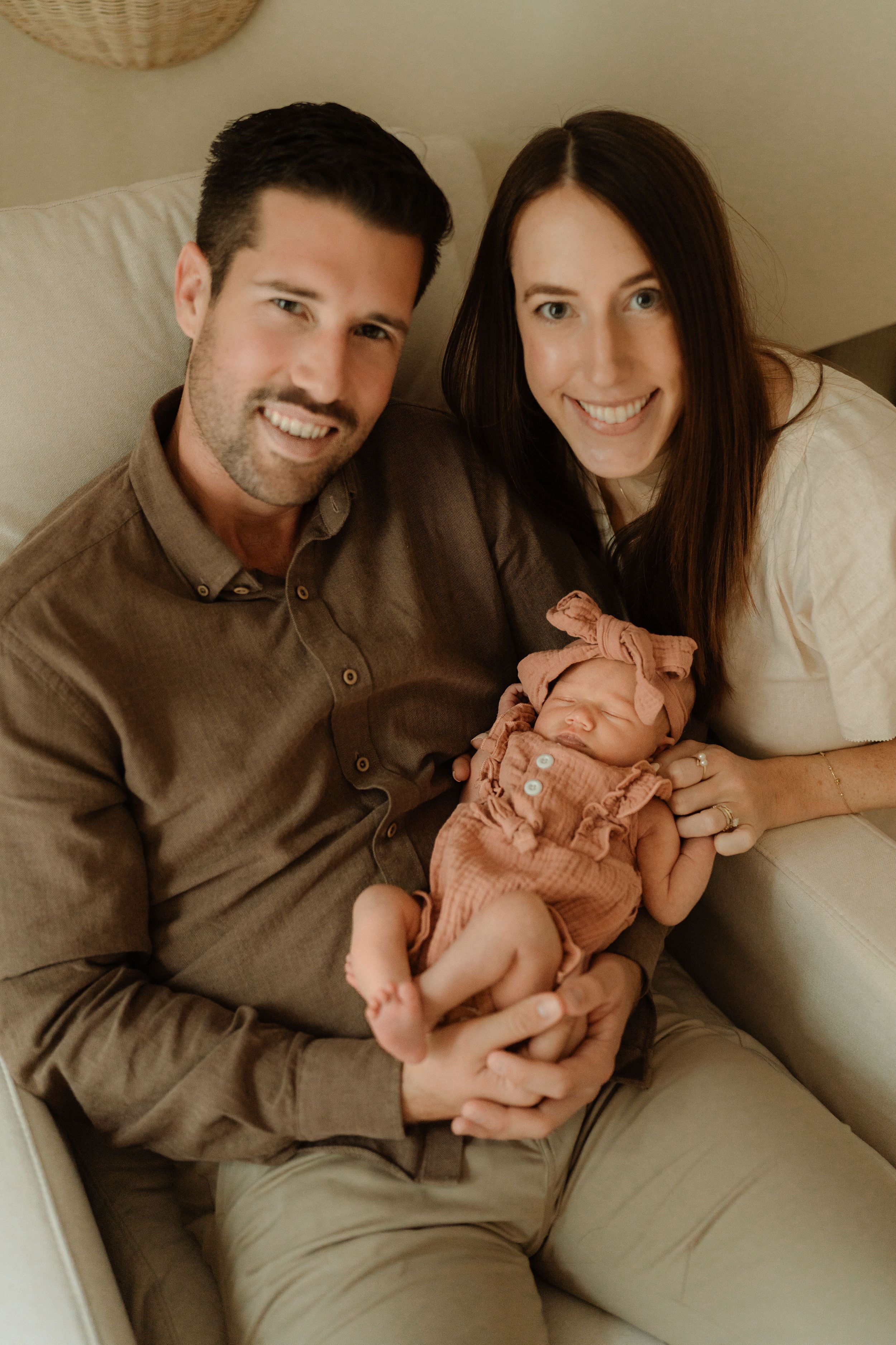 Smiling man and woman sitting together on a cream couch, gently holding a sleeping newborn baby wrapped in a soft peach romper.