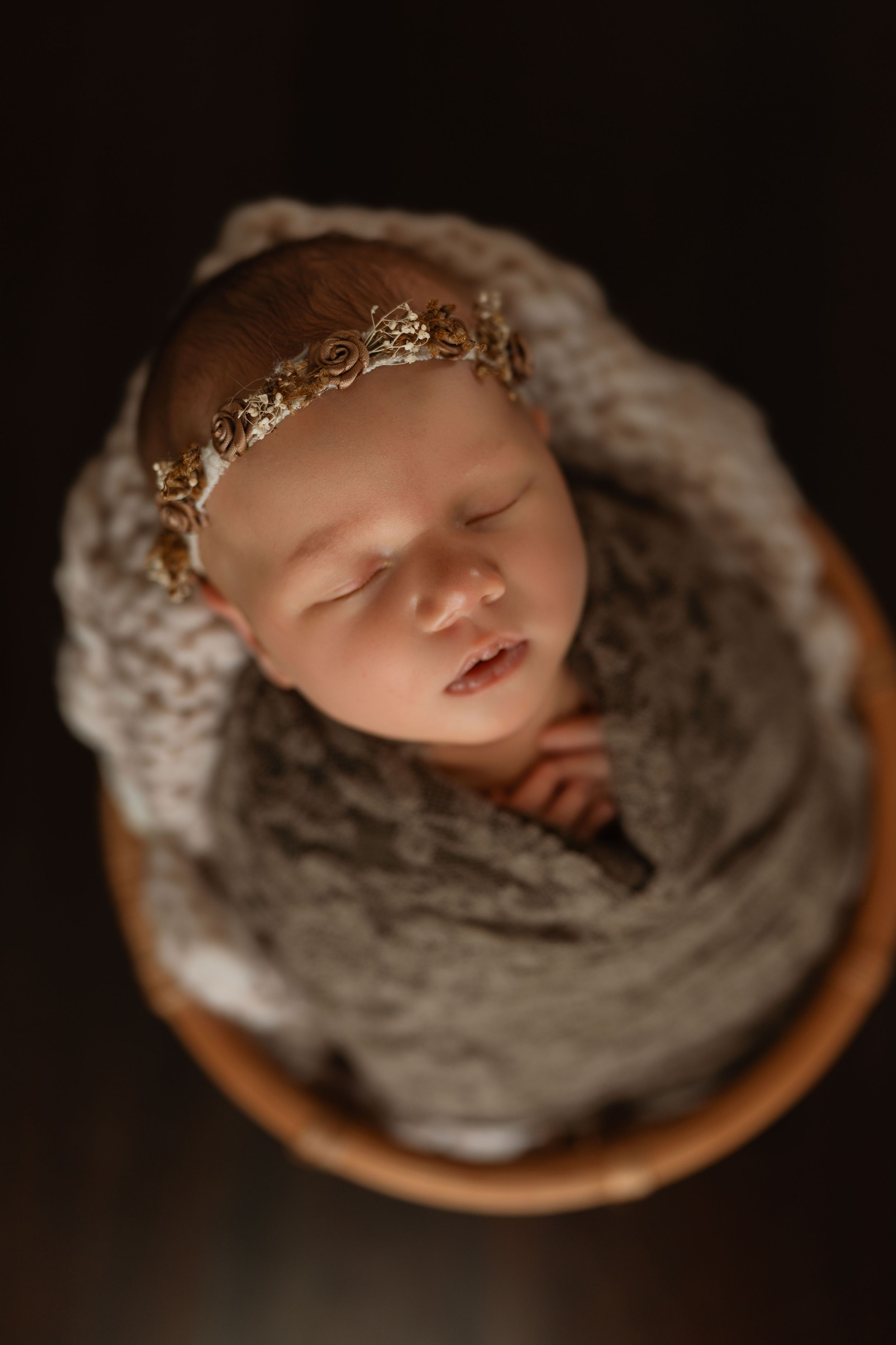 Sleeping newborn baby wearing a gold floral headband, swaddled in a soft gray knit wrap and resting in a wooden bowl prop.