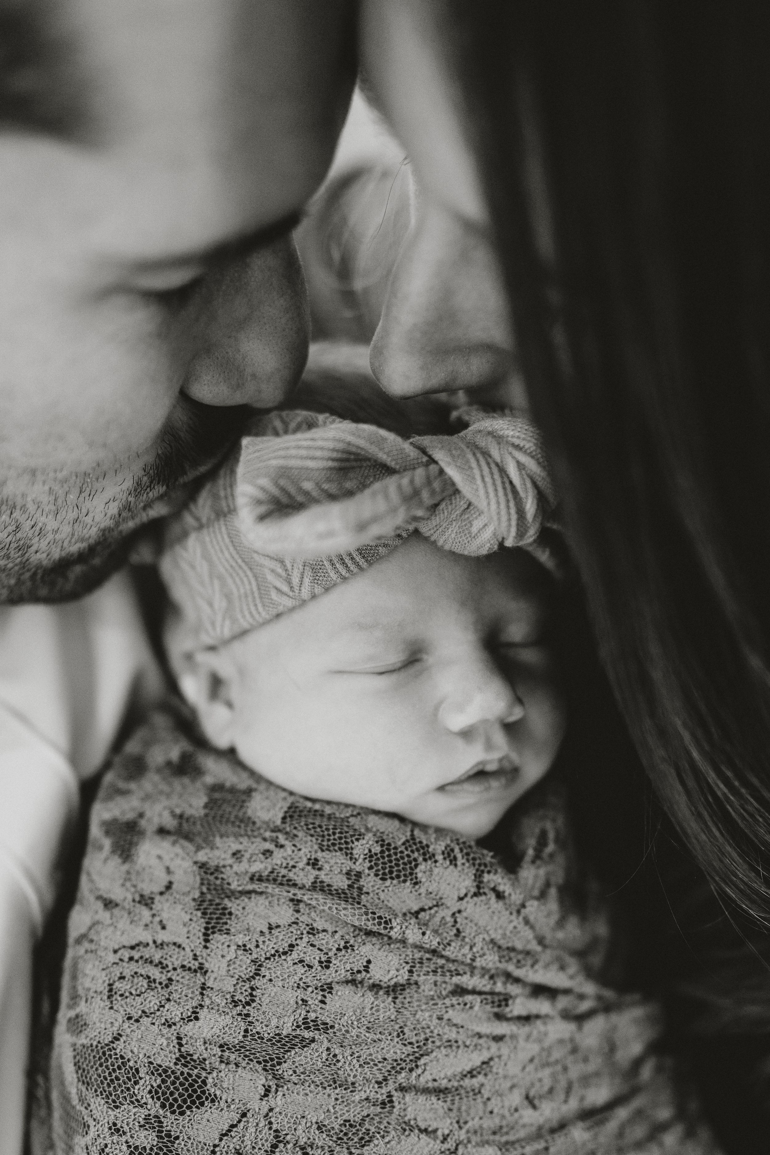 Black and white newborn photo of a sleeping baby in a knotted headband and lace wrap, nestled between two parents leaning in to kiss her.
