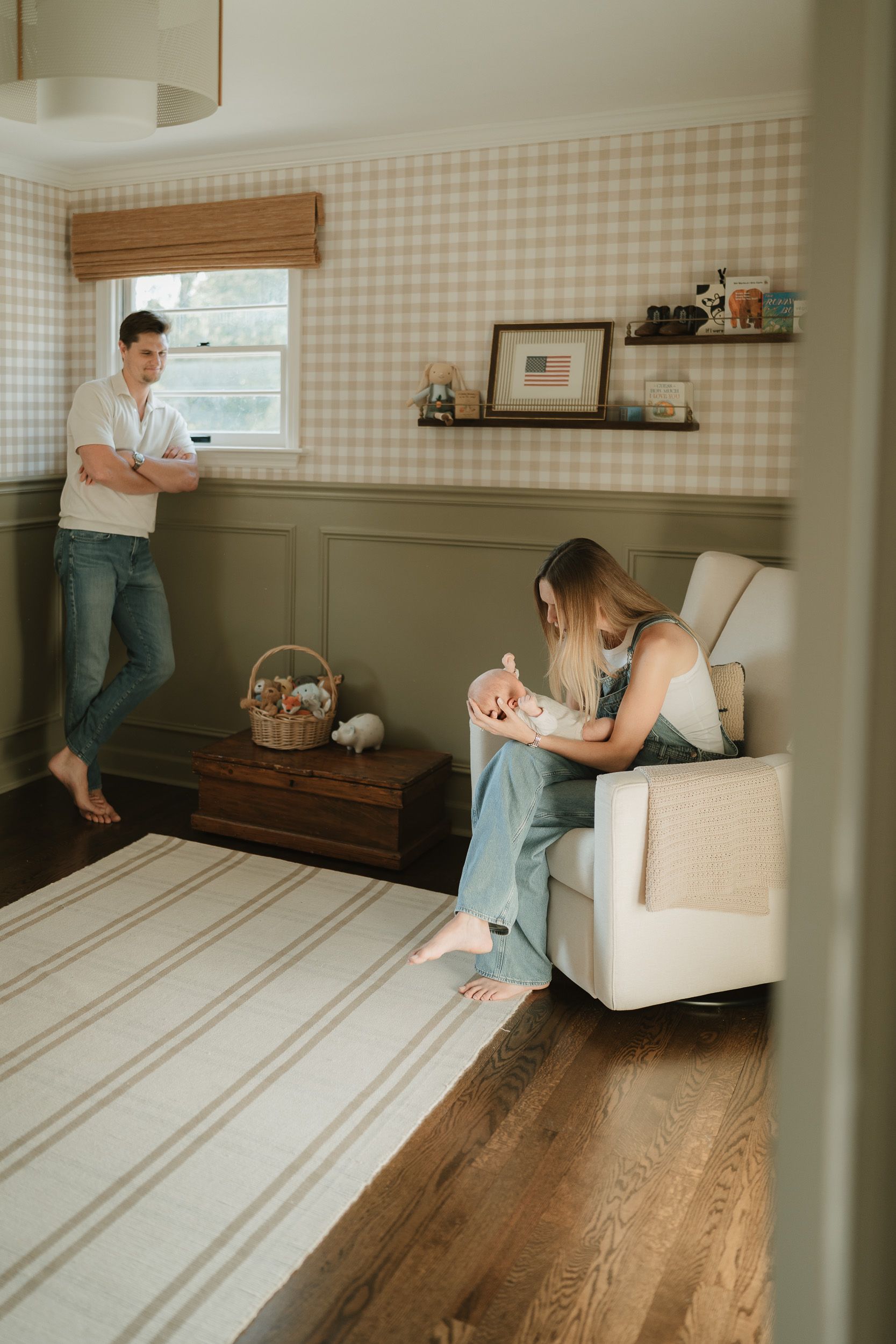 New parents in a cozy nursery with gingham wallpaper, mother holding newborn in glider chair, father standing nearby.