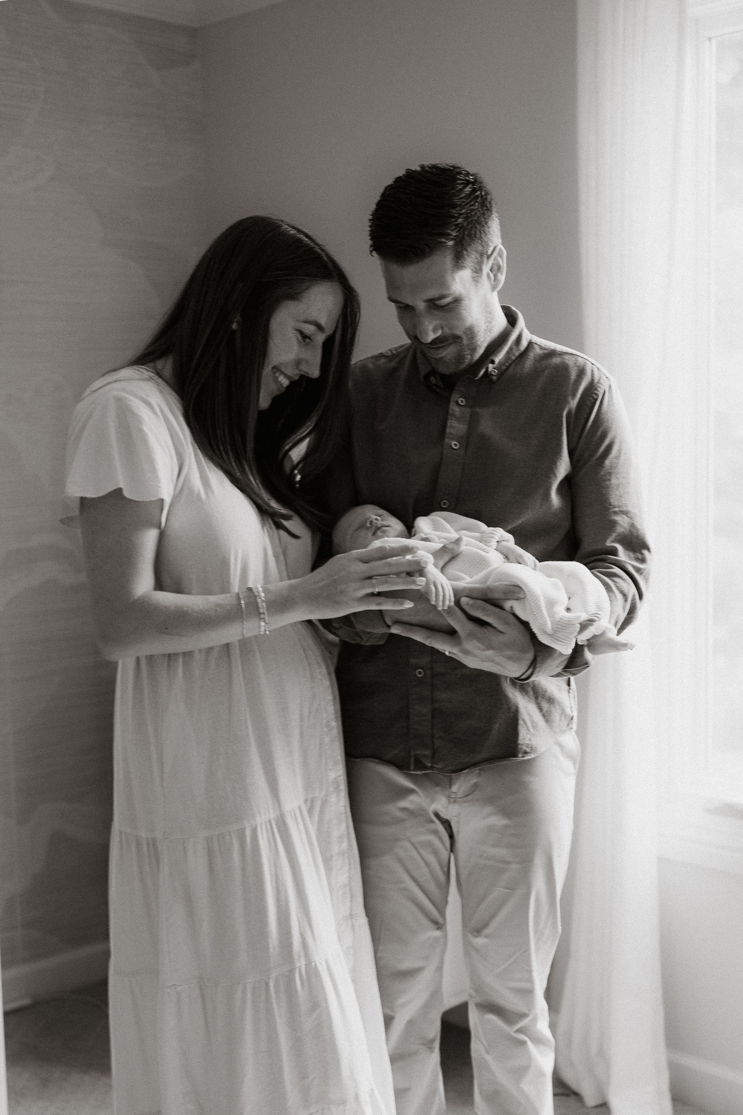 Black and white newborn photo of smiling parents standing by a window, gazing lovingly at their sleeping baby wrapped in a soft blanket.