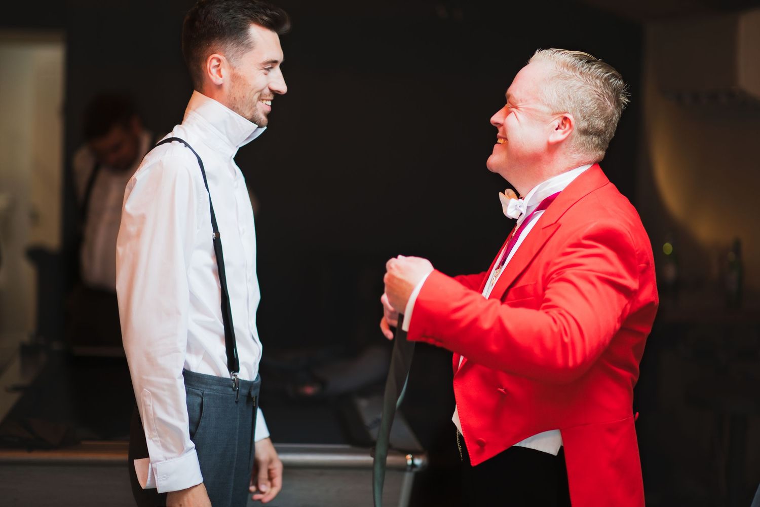 Neil, master of ceremonies at Merrydale Manor, in his red coat helping a groomsman with his tie, both smiling