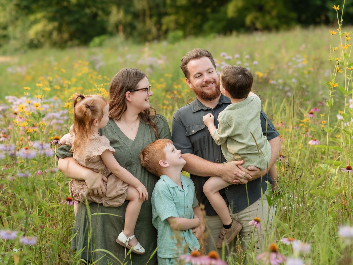 Happy family of four poses in a colorful wildflower meadow, parents holding young children while smiling together.