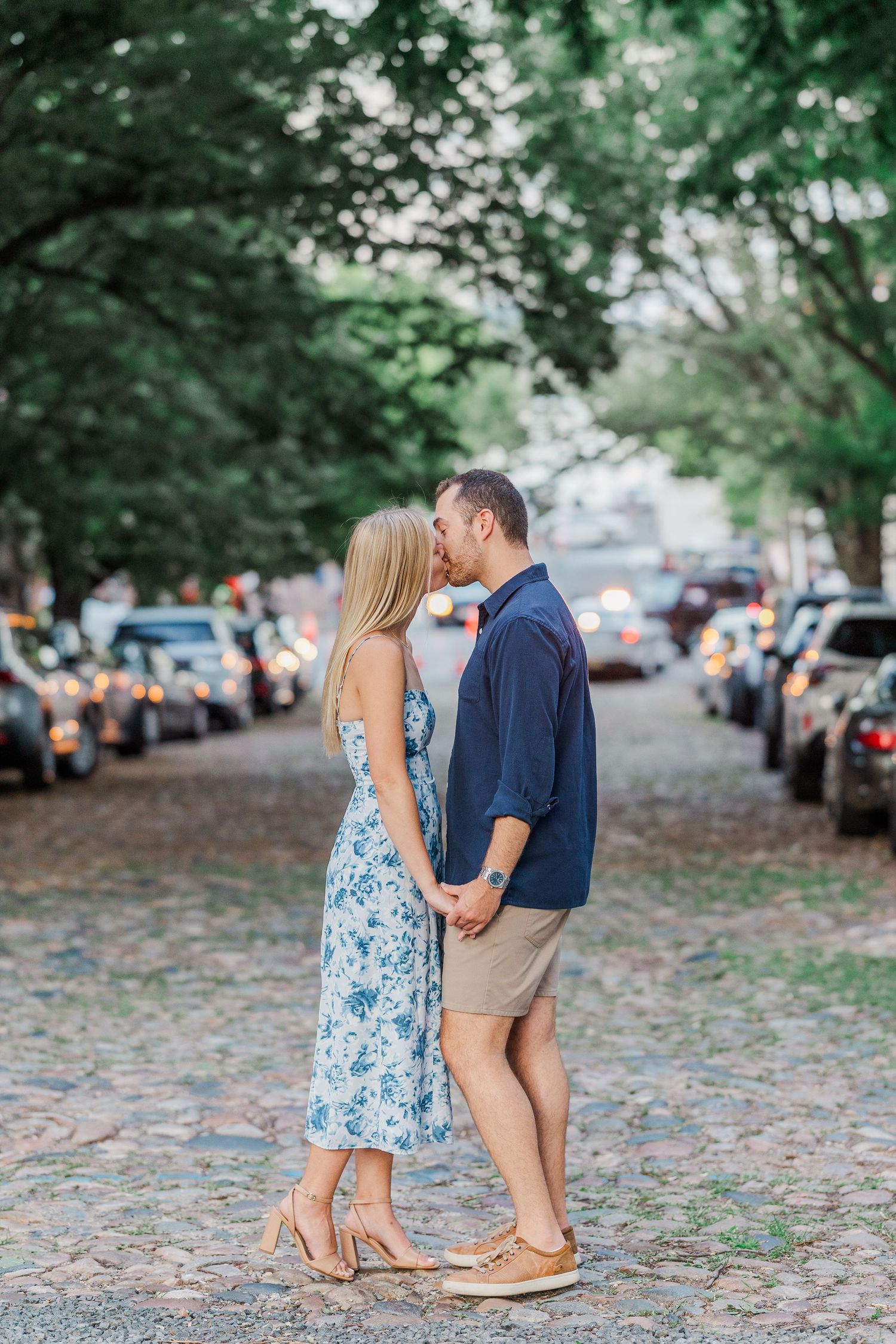 Couple sharing a romantic kiss on a cobblestone street lined with trees and parked cars, woman in floral dress.