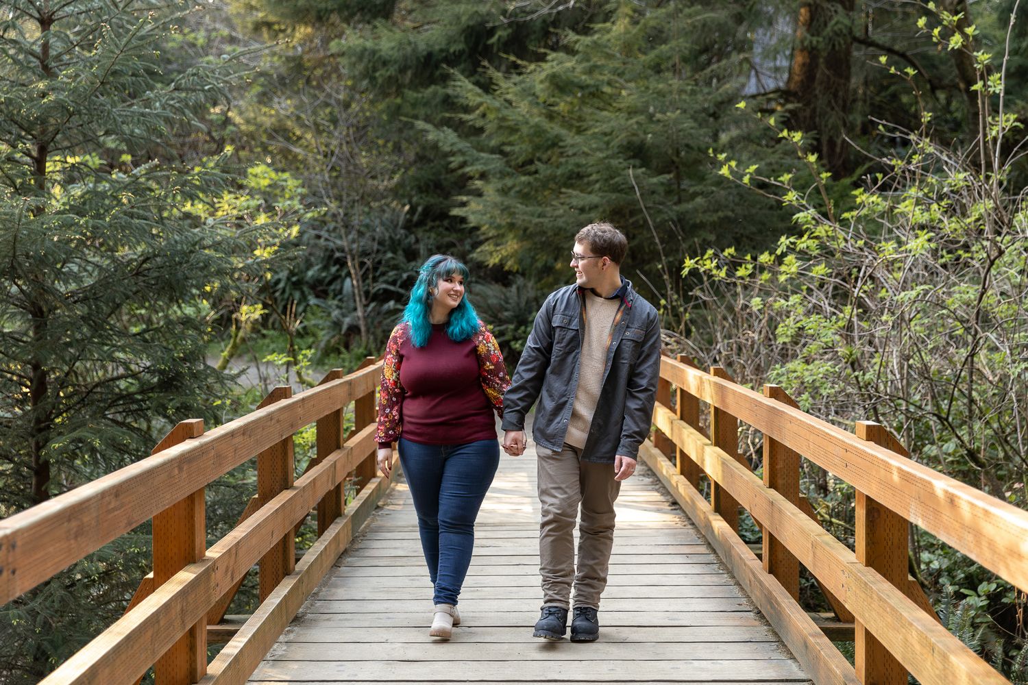 Couple holding hands walking on wooden bridge surrounded by lush green forest trees.