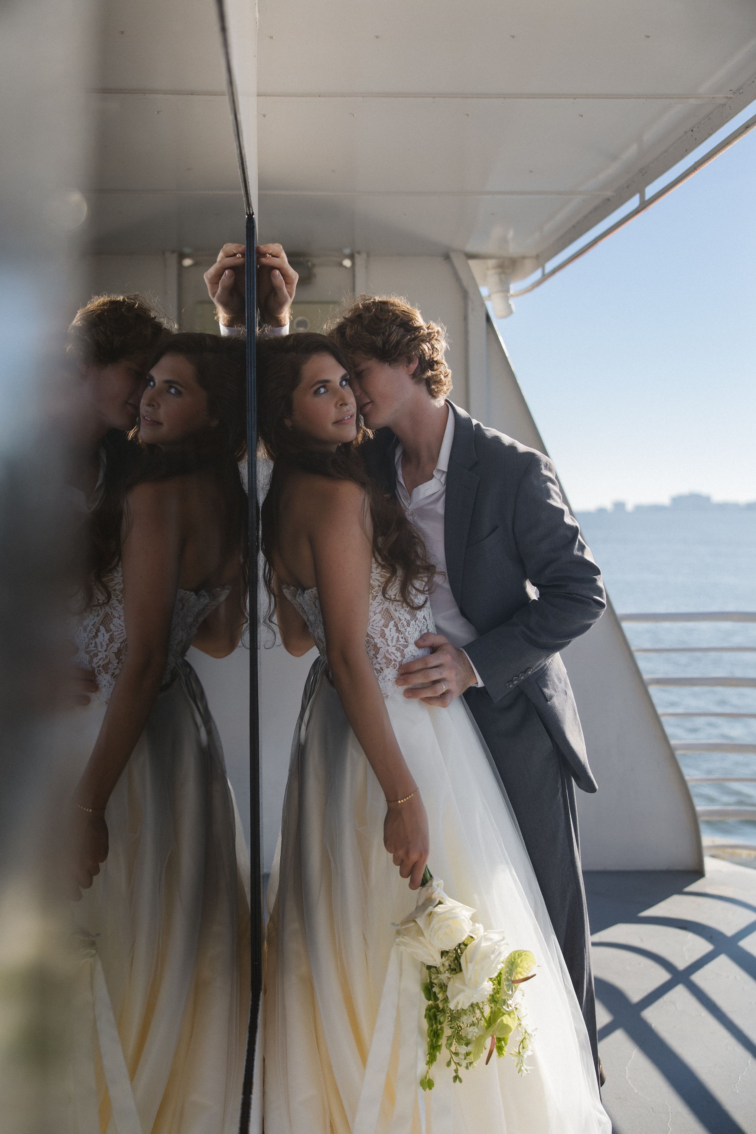 A bride in a lace wedding gown holding a white bouquet is kissed by her groom in a gray suit, reflected in a mirror on a yacht deck.