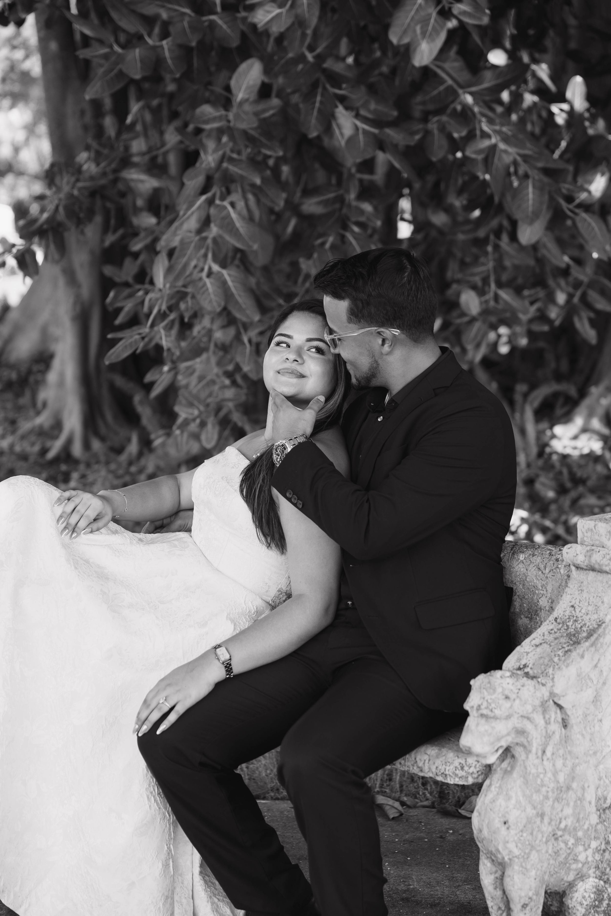 Black and white wedding photo of bride and groom sitting on stone bench under trees, sharing an intimate romantic moment.