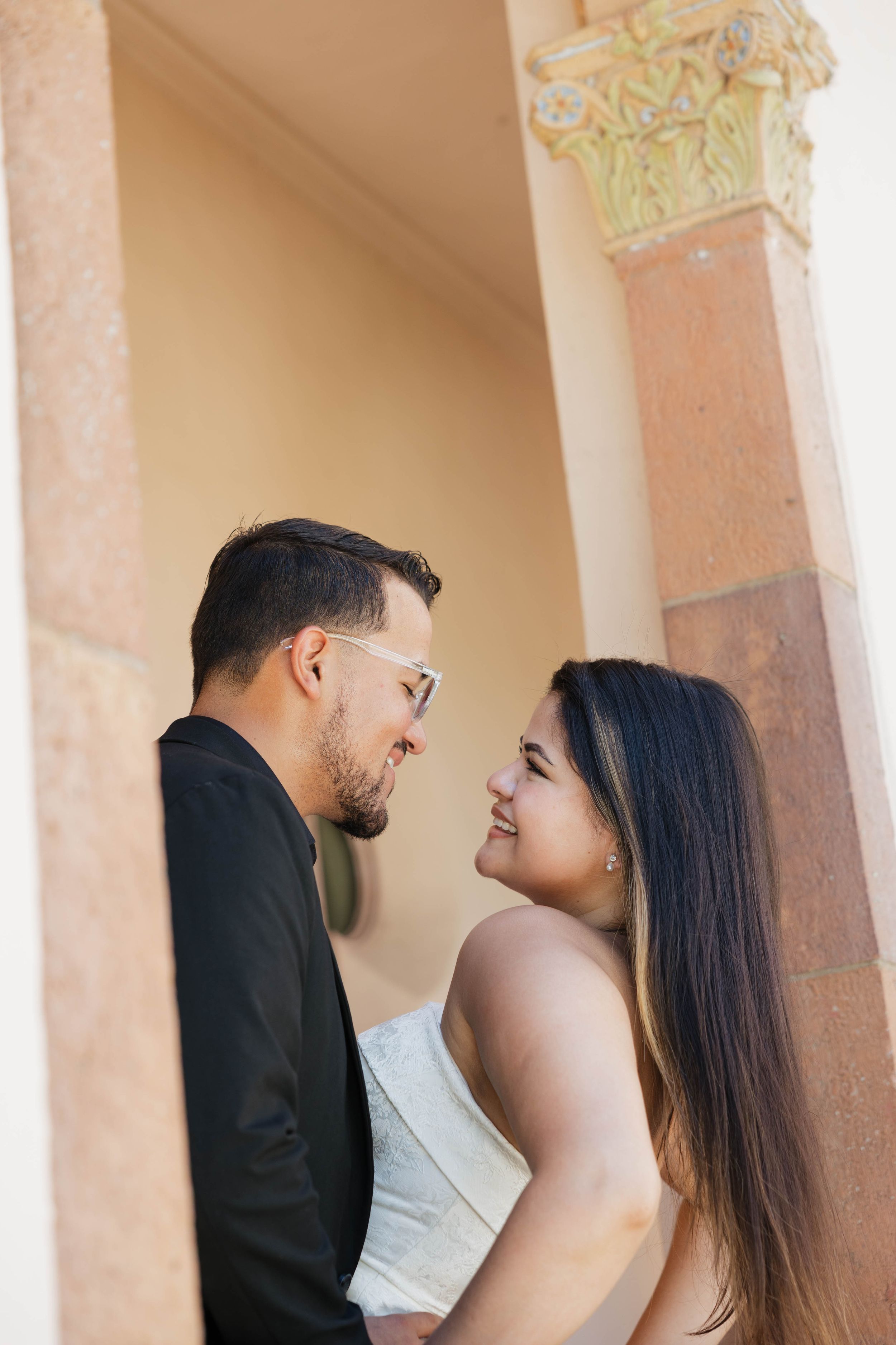 A couple sharing a romantic moment, smiling at each other near elegant stone columns in warm natural light.
